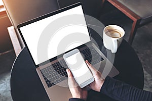 Mockup image of hands holding blank mobile phone while using laptop with blank white desktop screen on table