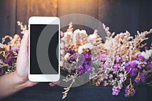 Hands holding and showing white mobile phone with blank black screen with colorful dry flowers and wooden wall background in cafe