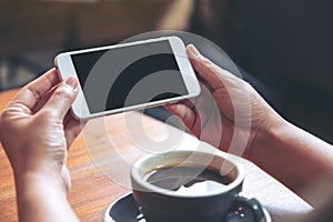 Mockup image of hands holding and using a white mobile phone with blank black screen horizontally for watching with coffee cup