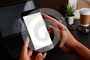 Mockup image of hands holding white mobile phone with blank black desktop screen and a coffee cup on desk