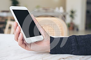 A hands holding white mobile phone with blank black desktop screen on table in modern cafe