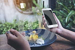 Mockup image of hands holding white mobile phone with blank black screen while eating yellow lemon curd cake on wooden table