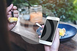 Mockup image of hands holding white mobile phone with blank black screen while eating yellow lemon curd cake on wooden table