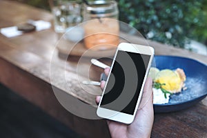 Mockup image of hands holding white mobile phone with blank black screen while eating yellow lemon curd cake on wooden table