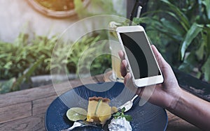 Mockup image of hands holding white mobile phone with blank black screen while eating yellow lemon curd cake on wooden table