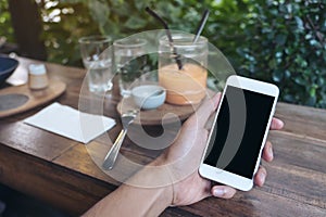 Mockup image of hands holding white mobile phone with blank black screen while eating yellow lemon curd cake on wooden table