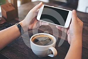 Mockup image of hands holding white mobile phone with blank black screen for watching and playing games with a cup of coffee