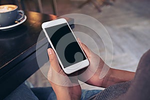 Hands holding white mobile phone with blank screen with coffee cup on table in modern cafe