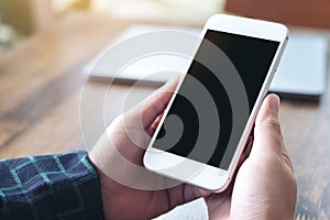 Mockup image of hands holding white smartphone with blank black desktop screen and laptop