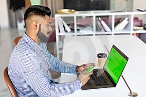 Mockup image of indian business man hand holding blank screen mobile smart phone while working on laptop computer in modern office