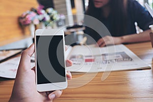 Mockup image of a man`s hand holding white mobile phone with blank black screen in modern cafe and blur woman reading newspaper