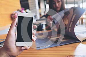 Mockup image of a man`s hand holding white mobile phone with blank black screen in modern cafe and blur woman reading newspaper