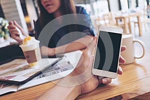 Mockup image of a man`s hand holding white mobile phone with blank black screen in modern cafe and blur woman reading newspaper