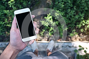 Mockup image of a man`s hand holding white mobile phone with blank black screen while standing on concrete floor with green nature