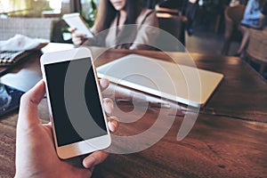 Mockup image of a man`s hand holding white mobile phone with blank black screen in vintage cafe with laptop on wooden table