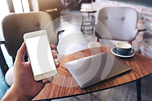 Mockup image of a man`s hands holding white mobile phone with blank desktop screen , laptop and coffee cup