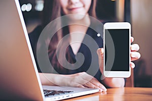 Mockup image of a smiley Asian beautiful woman holding and showing white mobile phone with blank black screen while using laptop