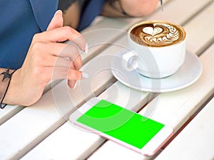 Mockup image of white mobile phone with blank black screen and hand holding hot latte coffee on vintage wood table in