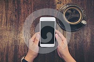 Mockup image of white mobile phone with blank black screen and hot coffee cup on vintage wood table background