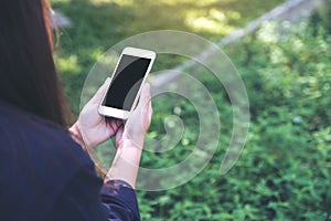 Mockup image of a woman holding and using white smart phone with blank black desktop screen in outdoor with green nature