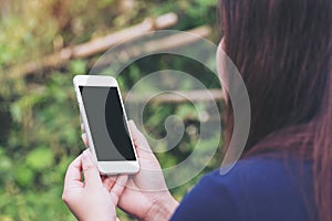 Mockup image of a woman holding and using white smart phone with blank black screen in outdoor and green nature