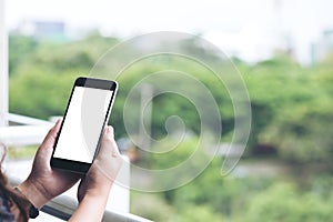 Mockup image of a woman`s hand holding and using black smart phone with blank white screen while standing at the balcony