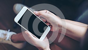 Mockup image of woman`s hand holding white mobile phone with blank black screen on thigh with wooden floor background