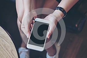 Mockup image of woman`s hand holding white mobile phone with blank black screen on thigh with wooden floor background