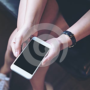 Mockup image of woman`s hand holding white mobile phone with blank black screen on thigh with wooden floor background