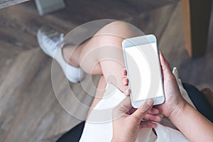 Mockup image of woman`s hand holding white mobile phone with blank screen on thigh with wooden floor background