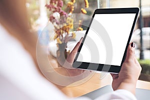 Woman`s hands holding black tablet pc with blank white desktop screen , laptop and coffee cup on wooden table in cafe