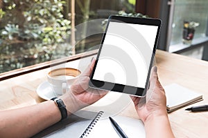 A woman`s hands holding black tablet pc with white blank screen with notebook and coffee cup on table