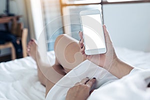 Mockup image of woman`s hands holding cell phone with blank desktop white screen while lying in a white bed