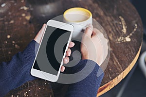 Mockup image of woman`s hands holding white mobile phone with blank black screen and a coffee cup on wooden table