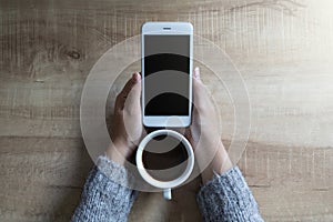 Mockup image of a woman`s hands holding white mobile phone with blank black screen with white hot coffee cups on wooden table in