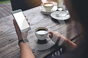 Mockup image of woman`s hands holding white mobile phone with blank screen and white coffee cup on wooden table