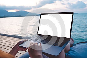 Mockup image of a woman using laptop with blank white desktop screen while sitting by the sea