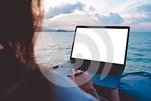 Mockup image of a woman using laptop with blank white desktop screen while sitting by the sea