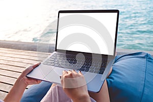 Mockup image of a woman using laptop with blank white desktop screen while sitting by the sea