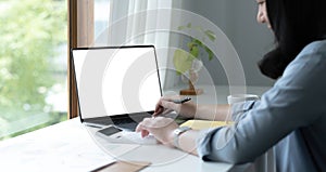 Mockup image of a woman using laptop with blank white screen on wooden table in office