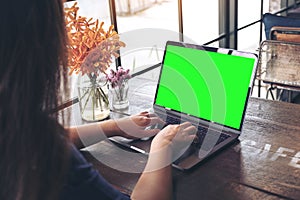 Mockup image of a woman using and typing on laptop with blank green screen and flower vase on vintage wooden table