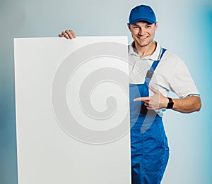 Mockup image of young smiling worker man wearing blue uniform. Holding white empty banner in his hand and pointing finger at you