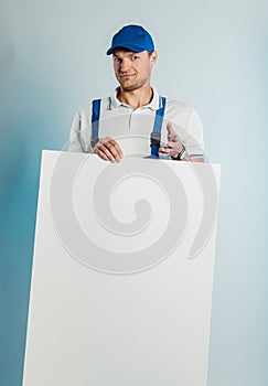 Mockup image of young smiling worker man wearing blue uniform. Holding white empty banner in his hand and pointing finger at you