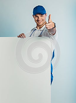 Mockup image of young smiling worker man wearing blue uniform. Holding white empty banner in his hand and pointing finger at you