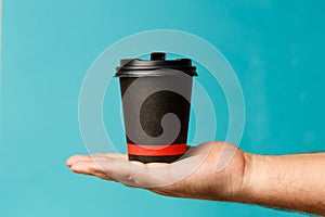 Mockup of man hand holding up a Coffee black color paper cup on blue background. Front view. Close-up of a hand