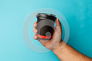 Mockup of man hand holding up a Coffee black color paper cup on blue background. Front view. Close-up of a hand