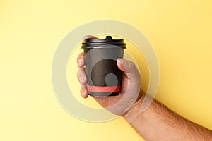 Mockup of man hand holding up a Coffee black color paper cup on yellow background. Front view. Close-up of a hand