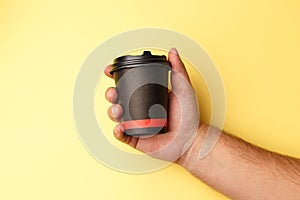 Mockup of man hand holding up a Coffee black color paper cup on yellow background. Front view. Close-up of a hand