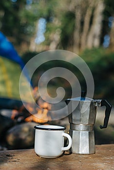 Mockup of metal camping aluminum cup or mug and coffee maker near bonfire on background tent. Concept adventures