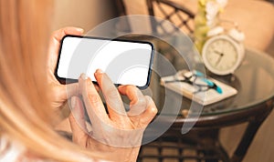 Mockup photo of woman holds smartphone horizontally with blank white screen, over shoulder view, home interior